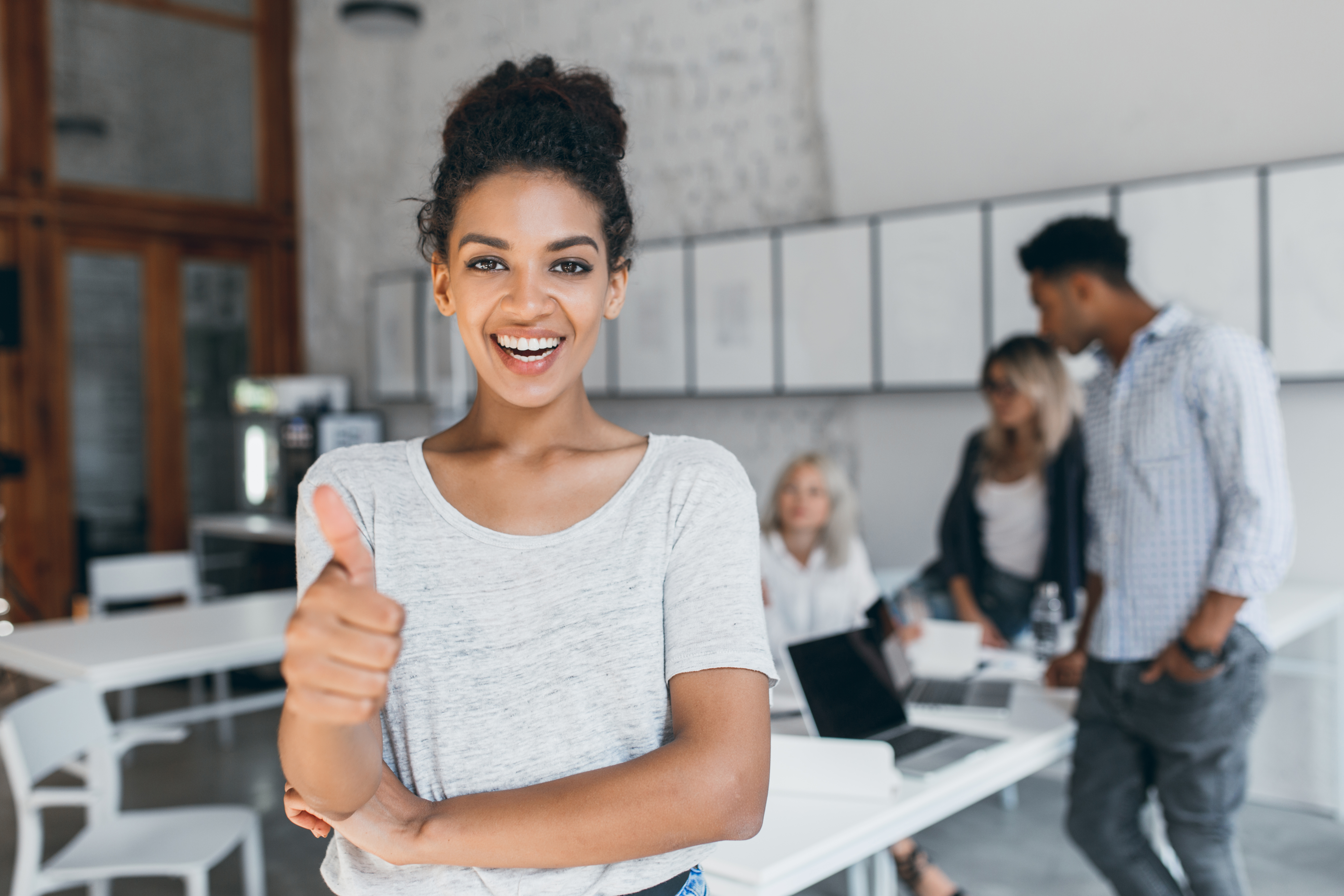 excited-mulatto-woman-with-curly-hairstyle-posing-with-thumb-up-office-stylish-female-african-student-having-fun-after-exams-with-university-friends